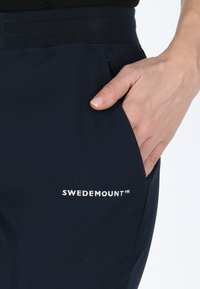 Pantaloni navy con cintura elastica, dotati di una tasca laterale. Logo "SWEDEMOUNT™" in bianco sul lato sinistro in basso. Texture liscia.