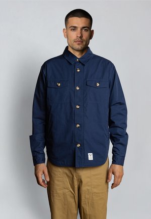 Mand iført en mørkeblå skjorte med knapper og brystlommer samt beige bukser, stående mod en ensartet lys grå baggrund.