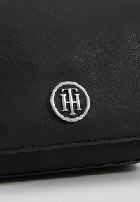 Černý texturovaný kožený povrch s malým stříbrným kulatým logem Tommy Hilfiger, na kterém se prolínají písmena T a H.