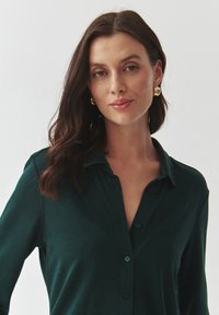 Zelená košile na knoflíky vyrobená z měkké tkaniny, s dlouhými rukávy a límečkem. Modelka má zlaté kruhové náušnice a lesklé rty.