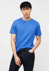 Νεαρός με κοντά μαύρα μαλλιά, φορώντας φωτεινό μπλε μπλουζάκι που είναι μπηγμένο μέσα σε σκούρα τζιν με μαύρη ζώνη, στέκεται μπροστά σε λευκό φόντο.