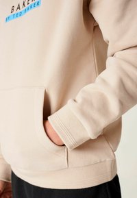 Sudadera beige con un logo azul, que presenta un bolsillo tipo canguro y puños acanalados. Textura suave, corte relajado y detalles de diseño minimalistas.