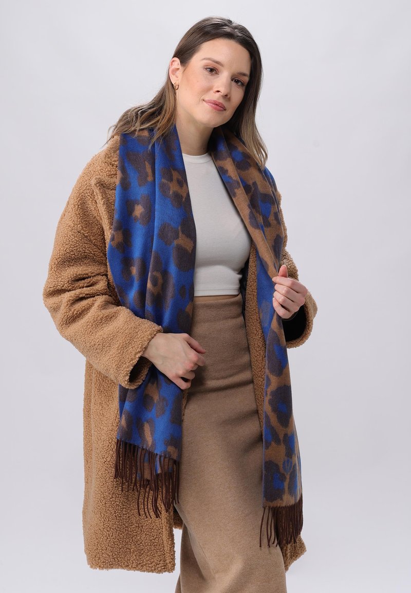 Abrigo de peluche marrón, falda beige, blusa blanca y una bufanda azul con estampado de leopardo marrón y flecos marrones, llevada de forma casual.