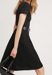 Svart, texturerad A-linjeformad klänning med korta ärmar, åtdragen i midjan med ett svart bälte, tillsammans med en svart handväska.