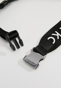 Czarny materiałowy pasek z metalowymi i plastikowymi elementami klamry, z logo „Michael Kors” na metalowym zapięciu.