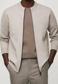 Cárdigan beige claro con cremallera y cuello redondo, combinado con una camisa beige. Presenta puños acanalados y un diseño elegante.