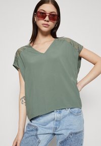 Top verde a V con maniche impreziosite da pizzo; tessuto morbido e leggero; vestibilità rilassata; indossato con jeans in denim a vita alta.