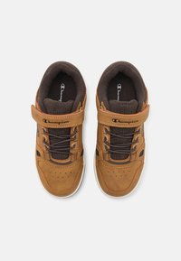 Zapatillas marrones con un upper texturizado, cordones negros y una correa de Velcro. Presenta acentos naranjas y perforaciones en la zona de los dedos.