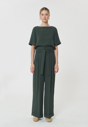 Femme portant un pantalon large vert foncé avec un haut assorti à manches courtes, debout devant un fond blanc uni.
