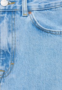 Gros plan sur un jean en denim bleu clair montrant la poche avant, la boucle de ceinture, le bouton et les détails de couture jaune.