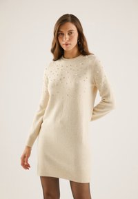 Light beige knitted sweater dress with a crew neck, embellished with metallic beads on the upper front, and ribbed cuffs and hem.