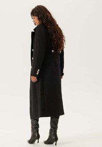Cappotto lungo nero realizzato in tessuto testurizzato, con bottoni dorati sulle maniche e sul retro. Abbinato a stivali neri al ginocchio.