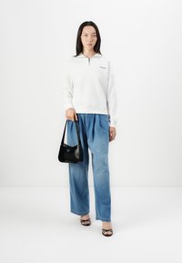 Felpa bianca con zip e logo, abbinata a pantaloni in denim blu larghi e una borsa a tracolla nera. Modella in sandali neri con i lacci.