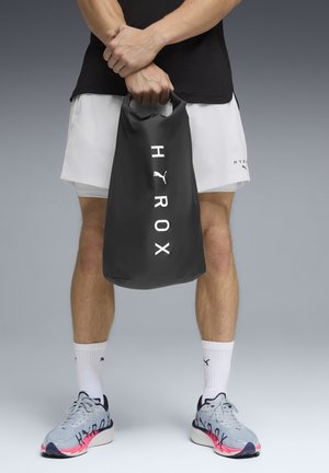 Persona que lleva pantalones cortos blancos y zapatillas grises sostiene una bolsa seca negra con los logos "HYROX" y Puma en la parte delantera, sobre un fondo neutro.