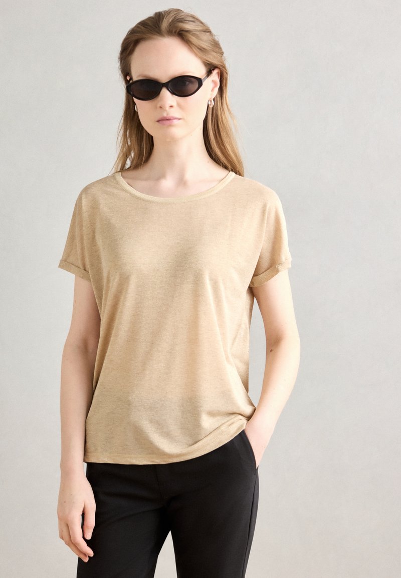 Beige t-shirt met korte mouwen in zachte stof, met een ronde hals. Combinatie met zwarte broek en kleine, ovale zwarte zonnebril.