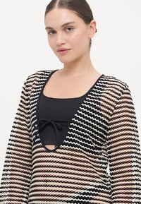 Blusa transparente de manga larga con un patrón zigzagueante en negro, blanco y óxido, con un escote en V profundo y detalle de cordón en el frente.