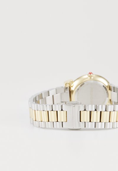 Montre en acier inoxydable avec un bracelet bicolore or et argent. Boîtier circulaire avec une touche de rouge sur la couronne. Texture métallique lisse.