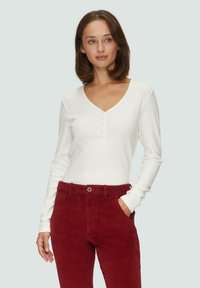 Weißes geripptes Langarmoberteil mit V-Ausschnitt und Knopfdetails, kombiniert mit hoch taillierten roten Cordhosen mit Fronttaschen.