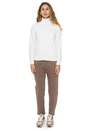 Una mujer está de pie sobre un fondo blanco y lleva una chaqueta blanca con cremallera, pantalones marrones y zapatillas deportivas beige.