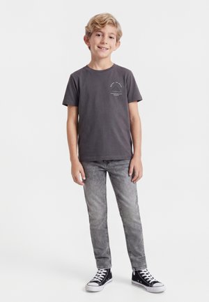 En ung dreng står og smiler, iført en mørkegrå t-shirt, grå jeans og sorte høje sneakers med hvide snørebånd.