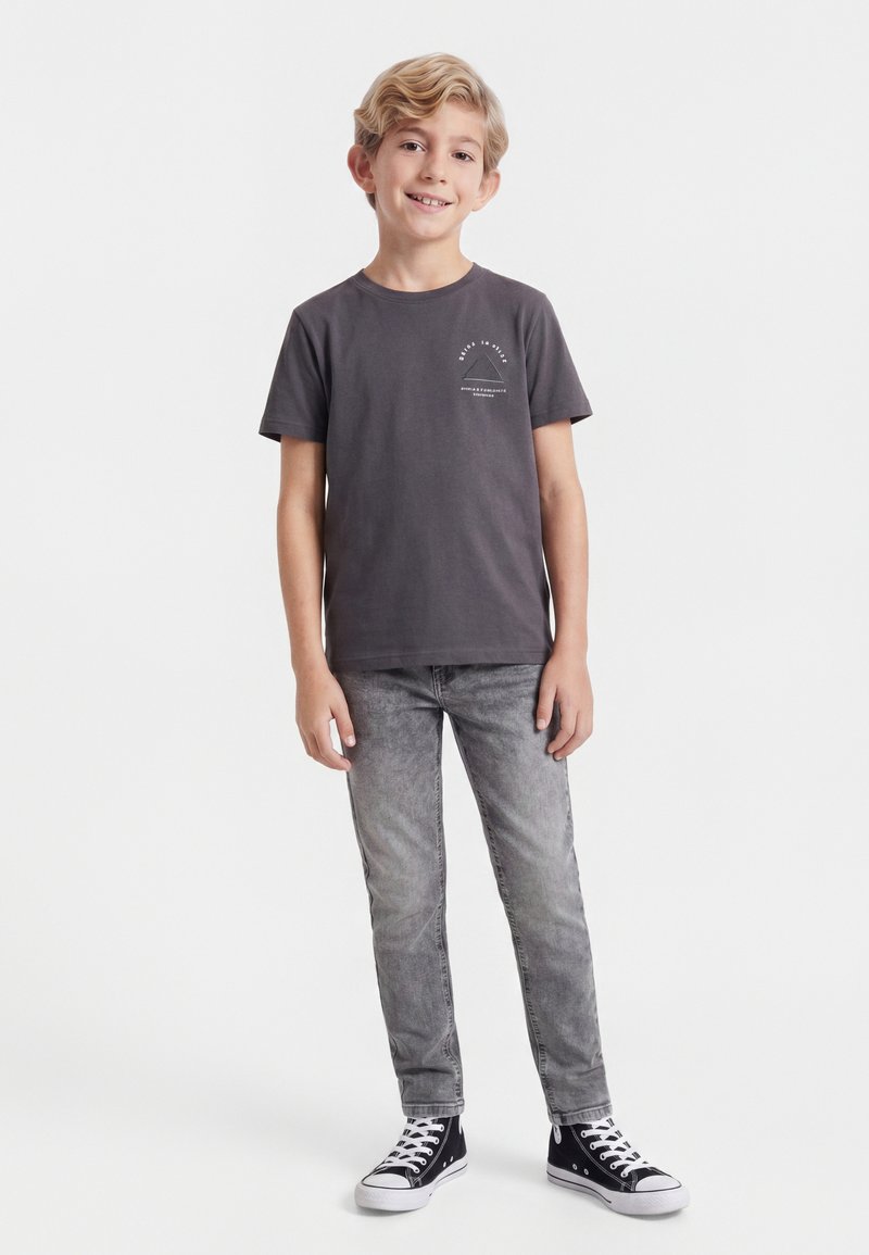 Jeune garçon debout et souriant, portant un t-shirt gris foncé, un jean gris et des baskets montantes noires avec des lacets blancs.