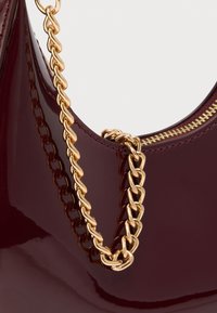 Sac à main brillant de couleur bordeaux foncé avec une bandoulière en chaîne dorée et une fermeture éclair dorée visible sur un fond clair uni.