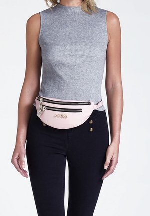 Ce sac banane en simili cuir rose possède des zips dorés, deux compartiments avant et le logo « GUESS » embossé. Porté à la taille sur un pantalon noir.