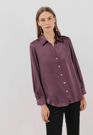 Halenka s dlouhým rukávem a límečkem v tmavě purpurové saténové úpravě, s knoflíkovou přední částí a perlou jako akcenty, zakřiveným lemem, kombinovaná s černými kalhotami.
