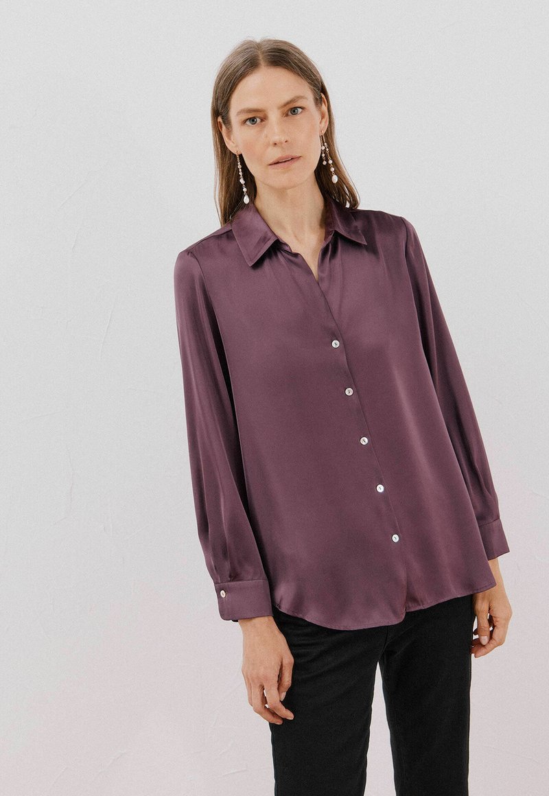 Blusa a maniche lunghe con colletto in satin viola scuro, con parte anteriore con bottoni e dettagli in perle, e orlo curvo, abbinata a pantaloni neri.