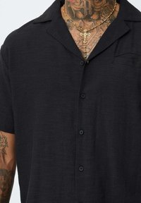Chemise noire à manches courtes avec un fini texturé, dotée d'un col en pointe et d'une seule poche poitrine. Des boutons noirs viennent parfaire le look.
