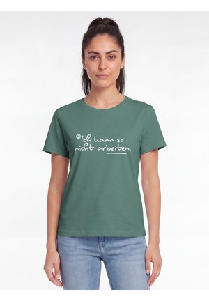Junge Frau mit dunkelbraunen Haaren im Pferdeschwanz, die ein grünes T-Shirt mit weißem deutschem Text "Ich kann so nicht arbeiten" und blaue Jeans trägt.
