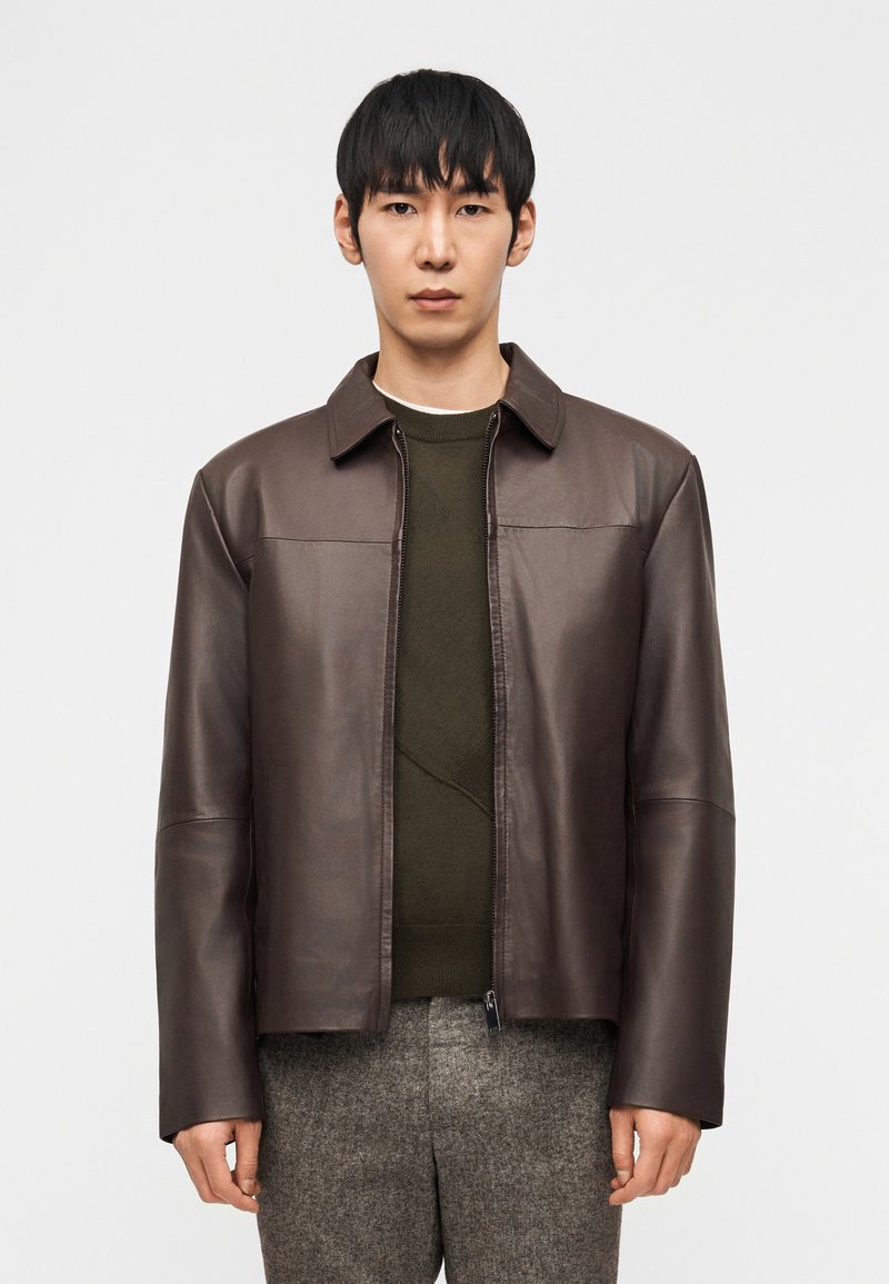 Άνδρας που φορά σκούρο καφέ δερμάτινο μπουφάν πάνω από ένα ελιά πράσινο πουλόβερ και γκρι μάλλινο παντελόνι, στέκεται μπροστά σε μια απλή λευκή φόντο.