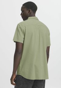 Camicia a maniche corte di colore verde oliva chiaro in un tessuto testurizzato con orlo curvo, dotata di colletto a punta e dettagli delle cuciture del yoke posteriore.