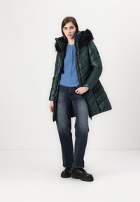 Cappotto puffer verde con collo in pelliccia sintetica nera, chiusura con zip e motivi a trapunta, abbinato a un maglione blu e jeans scuri. Scarpe nere chunky.
