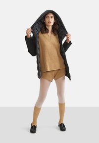 Zwarte pufferjack met een glanzende binnenkant, tan kleurige gebreide trui, bijpassende shorts en kniehooge beige sokken in combinatie met zwarte loafers.