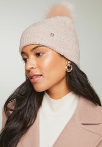 Căciulă tricotată de culoare roz deschis, cu textură ribbed, perle decorative și un pom-pom pufos pe vârf. Asortată cu un pulover bej cu guler înalt.