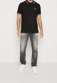 Camiseta de polo negra con cuello y puños a rayas rojas y azules, combinada con unos jeans gris claros y zapatillas blancas. Presenta un pequeño logo en el pecho.
