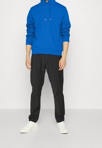 Uomo che indossa un felpa blu brillante, pantaloni neri a gamba dritta e scarpe da ginnastica bianche, in piedi contro uno sfondo chiaro.