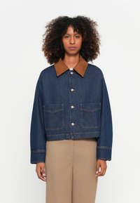 Donkerblauwe denim jacket met een bruine suède kraag, sluiting met knopen aan de voorkant en twee borstzakken. Heeft een cropped ontwerp en zichtbare stiksels.