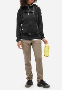 Schwarze Zip-Jacke mit Kapuze und Reißverschluss-Taschen, beige Stretchhosen, schwarze Wanderschuhe und eine gelbe Wasserflasche.