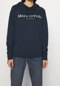 Granatowa bluza z kapturem ze sznurkiem, logo "Marc O'Polo" z przodu, ściągacze na mankietach i dole, wykonana z miękkiej bawełny.