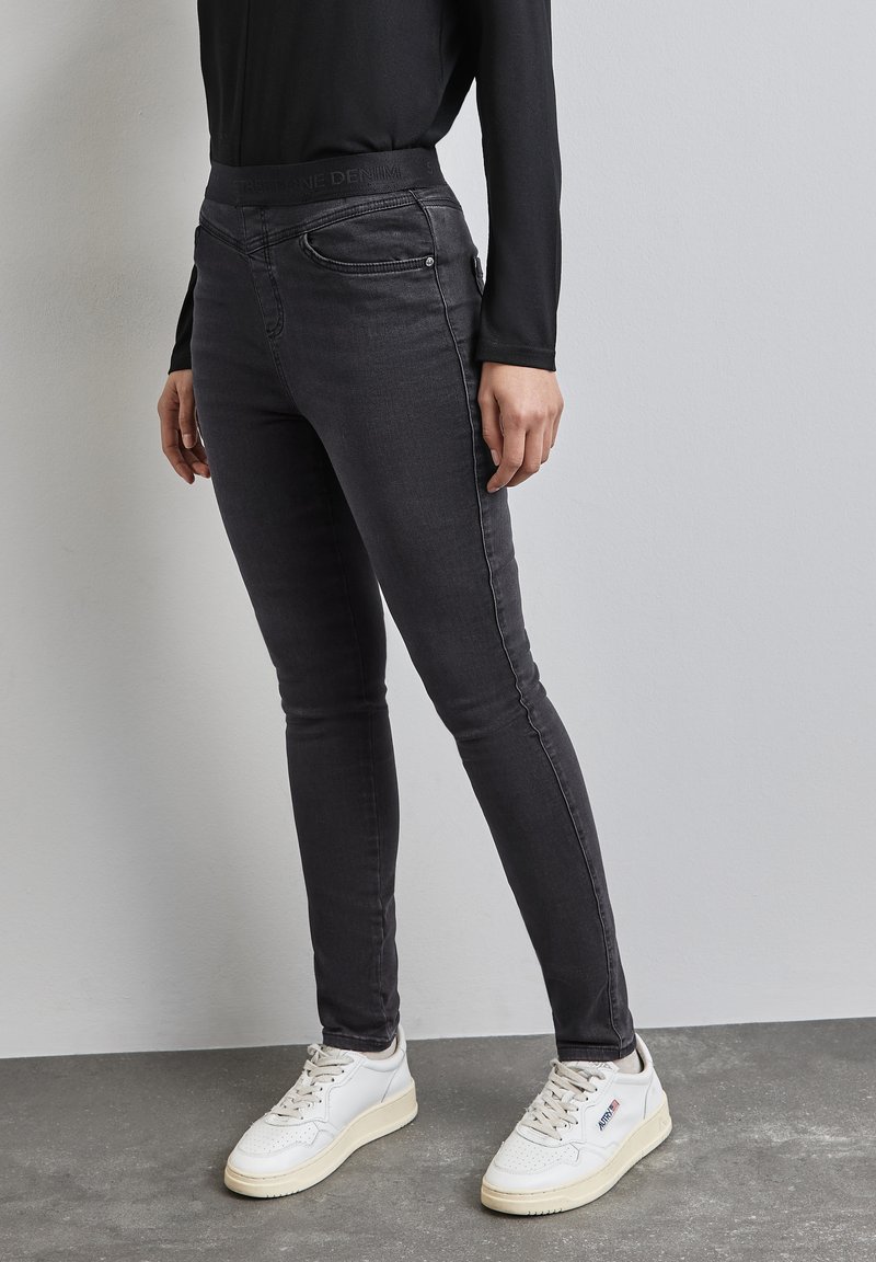 Slim-fit mustat farkut korkealla vyötäröllä, joissa on etutaskut ja sileä pinta. Yhdistettynä valkoisiin lenkkikengiin.