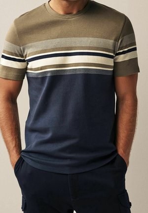 Homme portant un t-shirt à manches courtes col rond avec des rayures horizontales olive, blanc, marine et beige, associé à un pantalon foncé, mains dans les poches.
