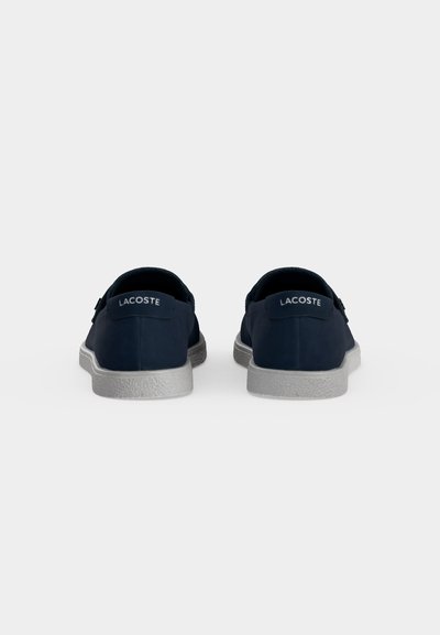 Двойка тъмносини спортни обувки Lacoste, показани отзад с текстурирани светлосиви подметки на плътен фон.