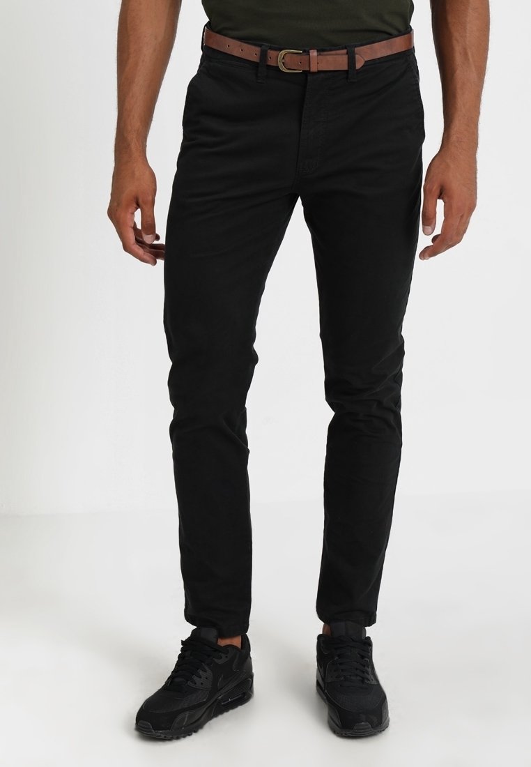 Homme portant un pantalon noir slim, une ceinture marron et des baskets noires, debout sur un fond blanc uni.