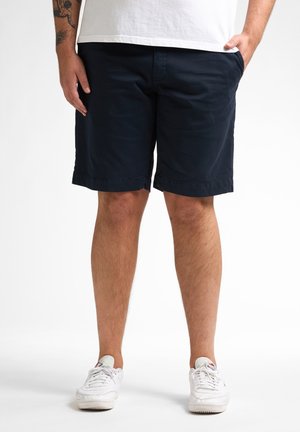Pantalones cortos azul marino de algodón, con corte recto, bolsillos laterales y dobladillo enrollado. Combinados con zapatillas blancas.
