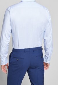 Camicia azzurra a maniche lunghe con una texture morbida, polsini abbottonati; abbinata a pantaloni blu scuri con tasche posteriori e dettagli con bottoni.