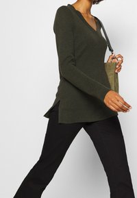 Maglione verde oliva a maniche lunghe con scollo a V, tessuto strutturato, spacchi laterali, abbinato a pantaloni neri attillati e una piccola borsa verde.