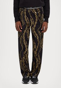 Black sweatpants with gold chain and logo pattern, featuring a relaxed fit and elastic waistband. Paired with black sneakers.