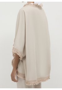 Beige poncho met franjes en een ruime pasvorm, gemaakt van een gestructureerde stof met een zachte afwerking, afgewerkt met franjes langs de mouwen en de zoom.